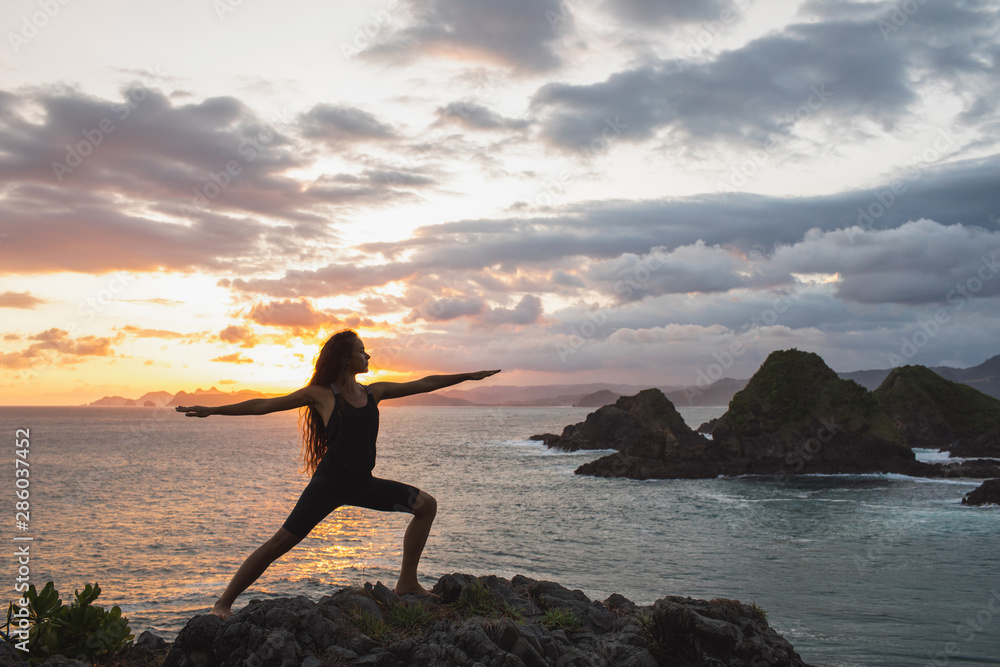 Young slim woman practicing yoga at sunset with beautiful ocean and mountain view. Workout outdoors, sport and well-being concept. Colorful and majestic sunset.
