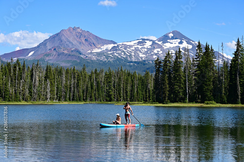 Young couple paddle boarding on Scott Lake, Oregon, with Middle and North Sisters volcanoes in the background on a calm sunny summer afternoon.