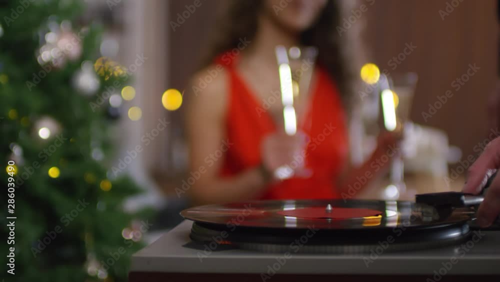 Panning close-up shot with bokeh of unrecognizable man in suit putting vinyl record on turntable, then joining girlfriend in red dress sitting with two glasses of champagne in blurred background Panning close-up shot with bokeh of unrecognizable man in suit putting vinyl record on turntable, then joining girlfriend in red dress sitting with two glasses of champagne in blurred background