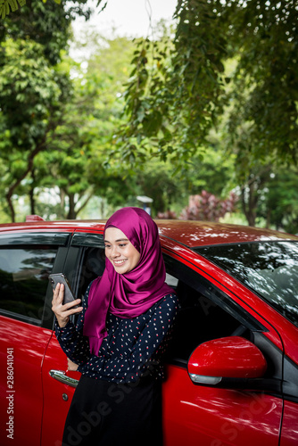 Happy Muslim Lady with her smartphone next to her car. Insurance Claim success concept.