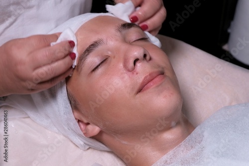 Face of a teenage boy with closed eyes. A beautician wipes a guy's cheeks and forehead with cotton pads. Dermatological procedure for cleansing teenager acne skin in a spa. Hygiene and personal care.