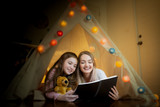 happy little girl and mother smiling and read book together in tents at home, creative festival thinking concept