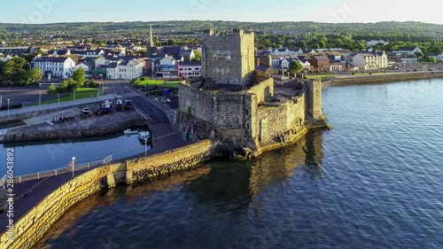 Belfast Lough with Medieval Norman Castle in Carrickfergus near Belfast in sunrise light. Aerial 4K flyby video with marina, breakwater, yachts, parking, town and far view of Belfast in the background