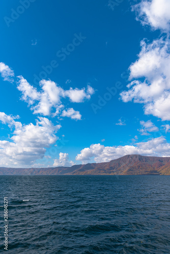 Beautiful autumn foliage scenery landscapes. Fall is full of magnificent colors. View from Lake Towada sightseeing Cruise ship. Clear blue sky, water, white cloud, sunny day background. Aomori, Japan © Shawn.ccf