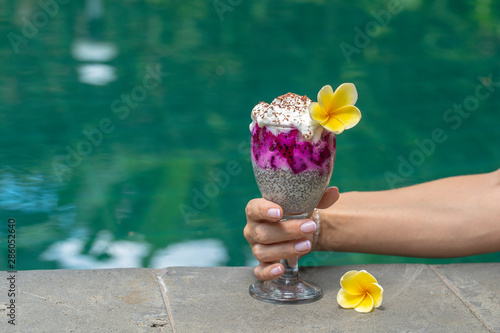 Girl hand holding a glass with hia seeds pudding on the background of the swimming pool water . Chia seeds pudding with red dragon fruit and white yogurt in a glass for breakfast photo