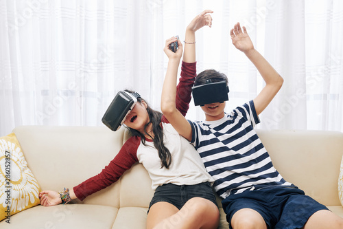 Teenage boy and girl sitting on sofa in living room and playing action virtual reality game