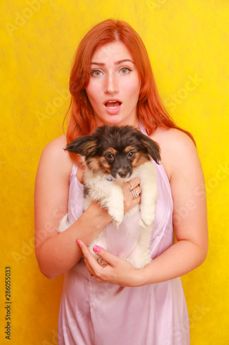 Relaxed red-haired girl embracing puppy on yellow background. Studio portrait of white appealing woman chilling with dog.