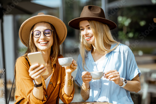 Two female best friends spending time together on the cafe terrace, feeling happy standing with coffee and phone during a summer day photo