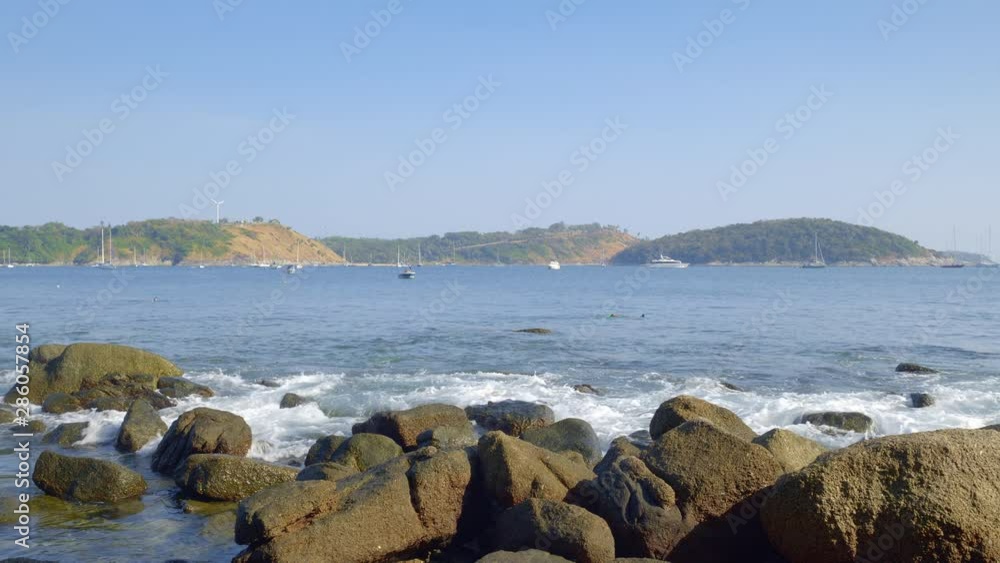 Rocky beach with many yachts, boats and catamarans, snorkeling people and paraglides in background over hill with wind power turbine Rocky beach with many yachts, boats and catamarans, snorkeling people and paraglides in background over hill with wind power turbine