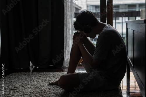 Wide shot young adult asian loneliness sad man sitting on the floor in bedroom leaning on bed and cry. Broken heart despair man sitting on the floor listening music from headphone alone.