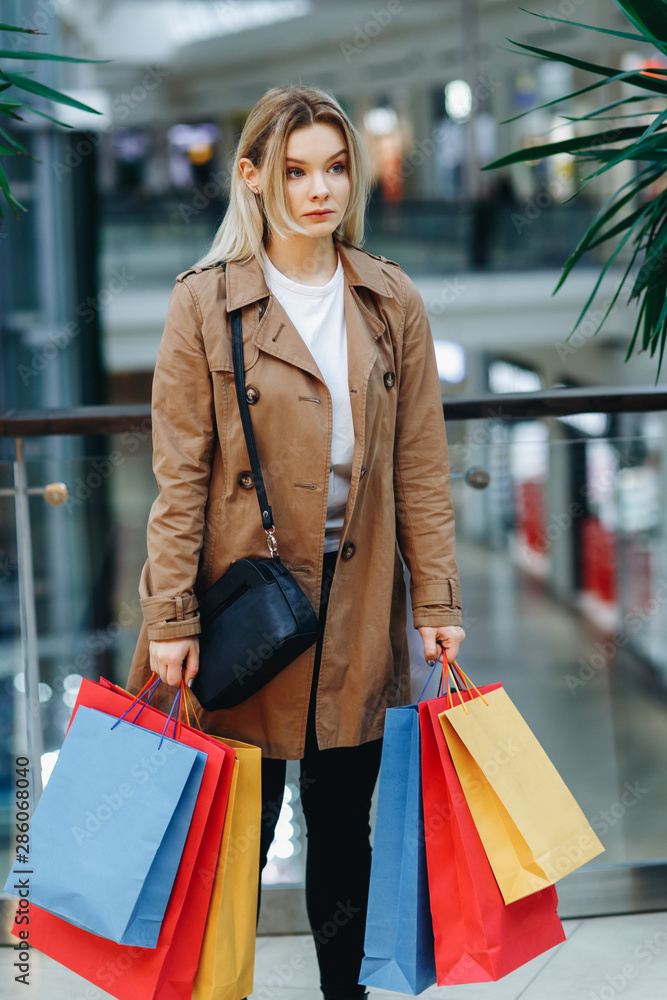 Woman after shopping. Serious young woman in a brown cloak at a shopping center on the background of boutiques. Beauty woman with colorful shopping bags in shopping mall Woman after shopping. Serious young woman in a brown cloak at a shopping center on the background of boutiques. Beauty woman with colorful shopping bags in shopping mall