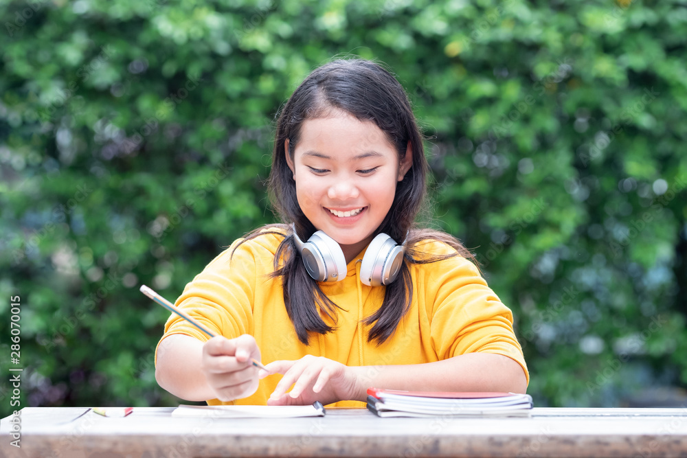Smiling little Asian schoolgirl doing her homework in the park with headphones on her neck Smiling little Asian schoolgirl doing her homework in the park with headphones on her neck