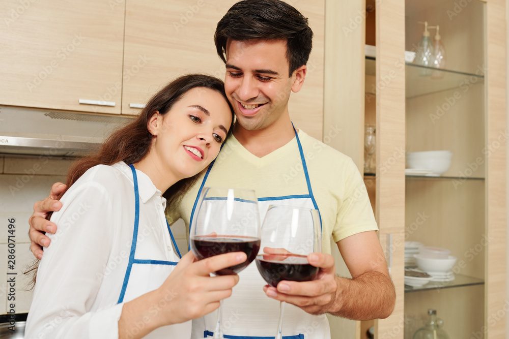 Positive young Caucasian couple in aprons standing in kitchen and drinking red wine after cooking together Positive young Caucasian couple in aprons standing in kitchen and drinking red wine after cooking together