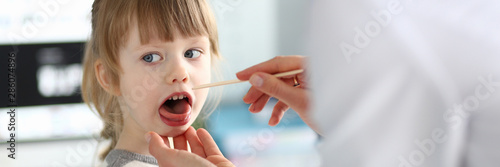 Female paediatrician examining little kid patient throat with wooden stick portrait