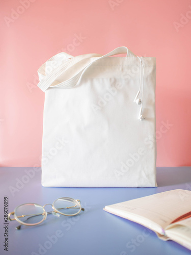 Mock up design bag concept. Blank white tote bag canvas fabric with earphones, notebook and glasses on pink purple background. Empty eco bag. Copy space. Vertical.