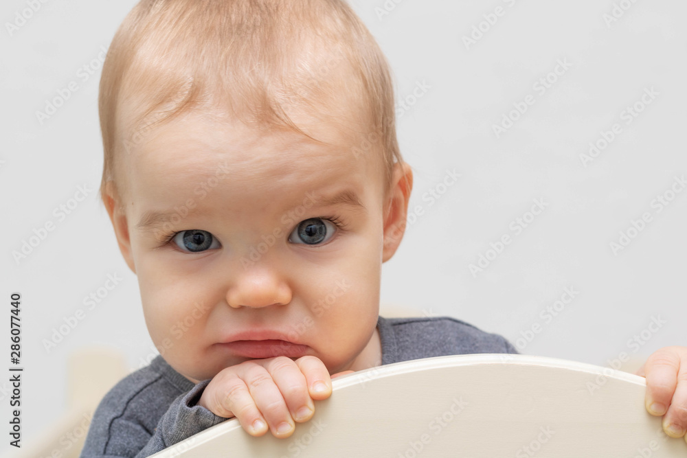 Portrait of a European Caucasian child baby sad sadly, wrinkles his forehead, looks at the camera, clings to the bed. On a white background. Age - 10 months - 1 year. Portrait of a European Caucasian child baby sad sadly, wrinkles his forehead, looks at the camera, clings to the bed. On a white background. Age - 10 months - 1 year.