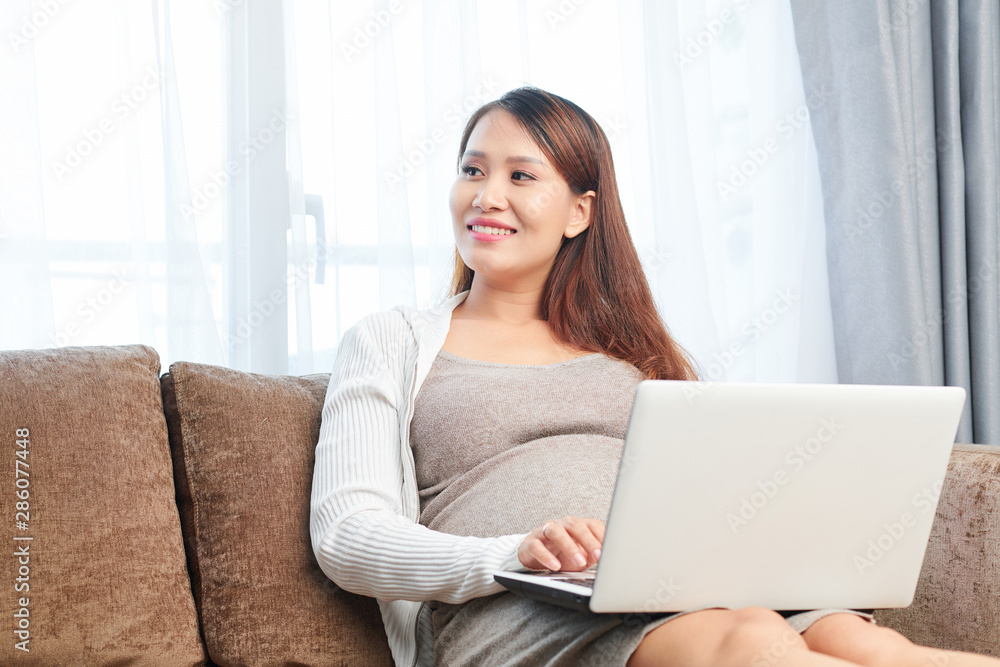 Smiling young pregnant Asian woman working on laptop when sitting on sofa at home Smiling young pregnant Asian woman working on laptop when sitting on sofa at home