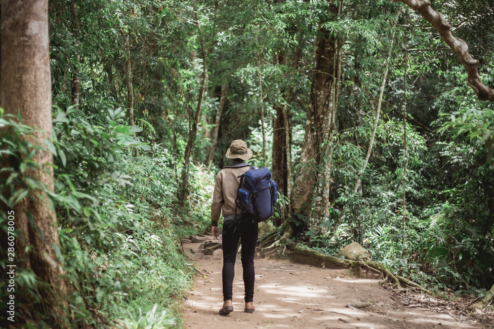 Fototapeta premium The back of an Asian young man carrying a suitcase and wearing a hat the front is a view of the forest and pond.