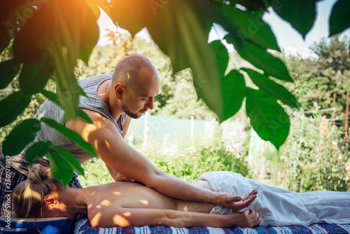 Female gets relaxing Ayurvedic massage in the open air among green trees. Young healthy woman enjoying the day at Spa, lying on the table during wellness procedure. Male back massage outdoor.