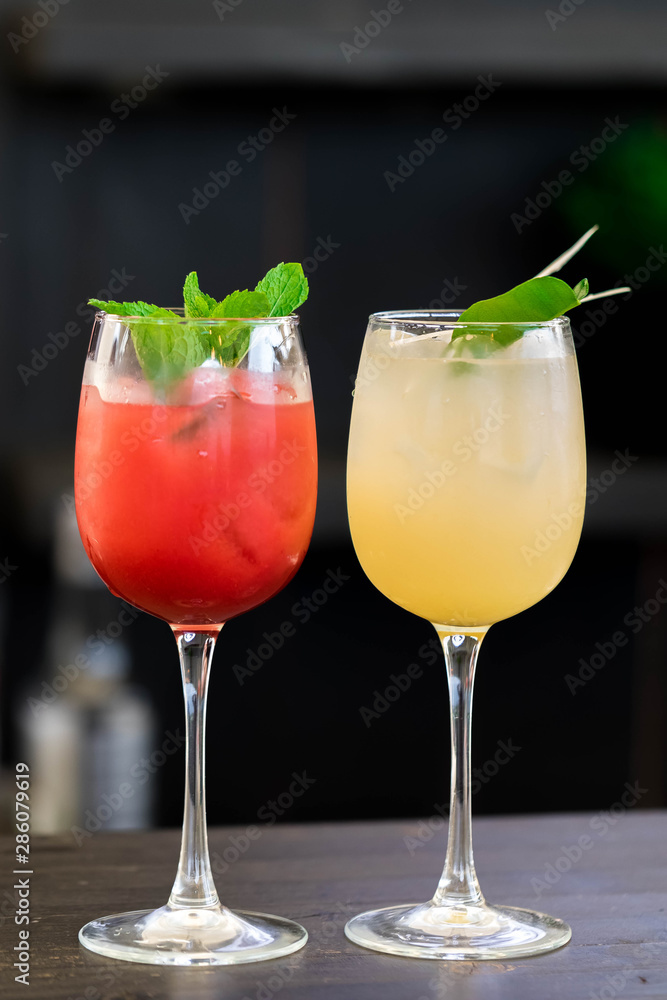 Two glasses with cocktails of the red and yellow colors decorated by leaves of fresh mint on a dark bar counter