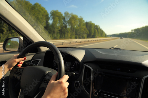 Female hands, lit by sunlight, hold the steering wheel of a car while driving on the road. © DredTorgal