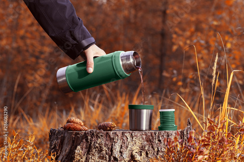 A man pours hot tea from a thermos into a mug on a stump. Autumn forest blurred