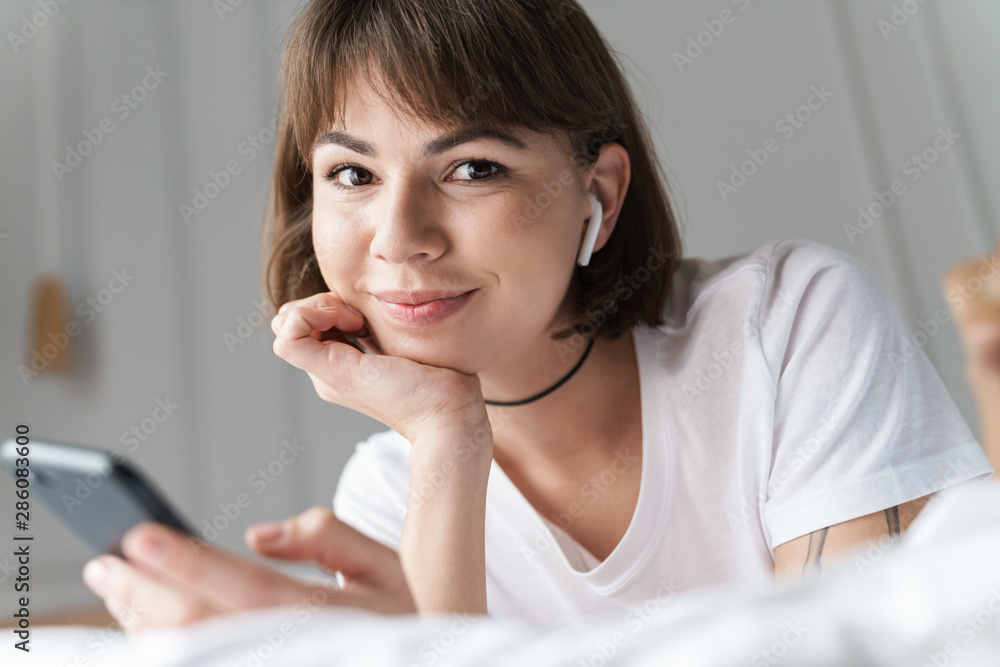 Smiling pleased young beautiful woman lies indoors at home on bed listening music with bluetooth earphones using mobile phone. Smiling pleased young beautiful woman lies indoors at home on bed listening music with bluetooth earphones using mobile phone.