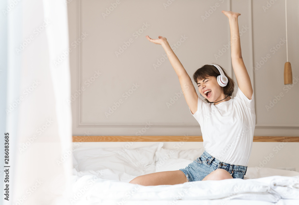 Cheery young beautiful woman sitting indoors at home on bed listening music with headphones.