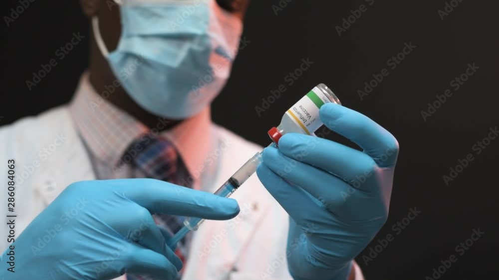 African American doctor drawing vaccine from vial into a syringe on a black background wearing white coat and blue gloves with mask African American doctor drawing vaccine from vial into a syringe on a black background wearing white coat and blue gloves with mask