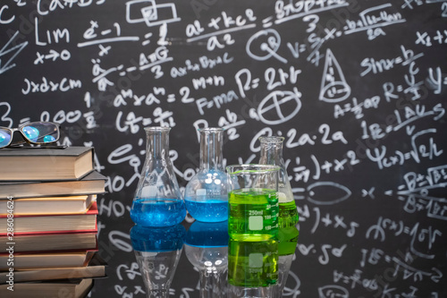 Glasses on old books and science experiment bottle on a black table with a blackboard on the background.