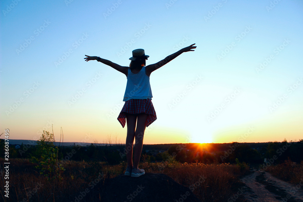 Silhouette of a young teenage girl  wearing a hat standing back, looking to  the sunset and holding hands up. People outdoors, travel, landscape concept. 