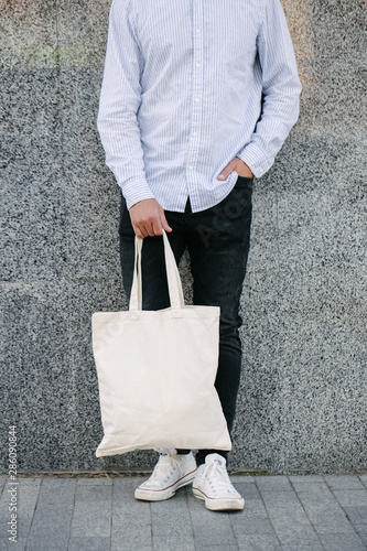 Young man holding white textile eco bag against urban city background. . Ecology or environment protection concept. White eco bag for mock up.