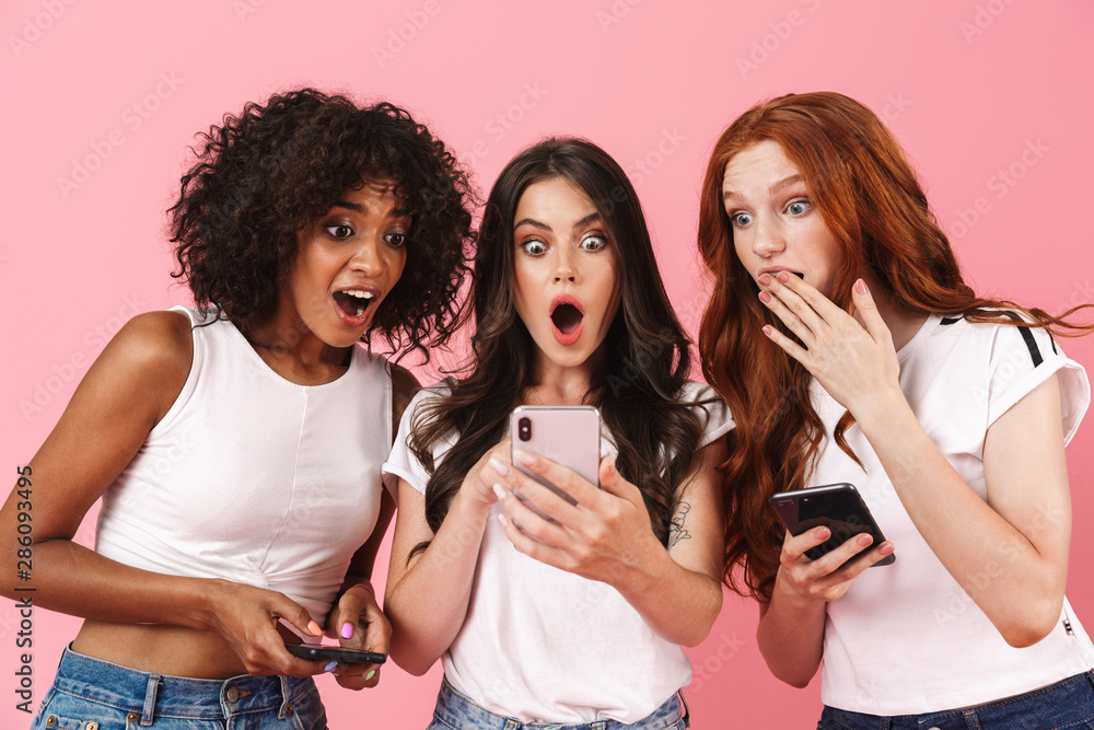 Surprised shocked emotional young three multiethnic girls friends posing isolated over pink wall background using mobile phones.