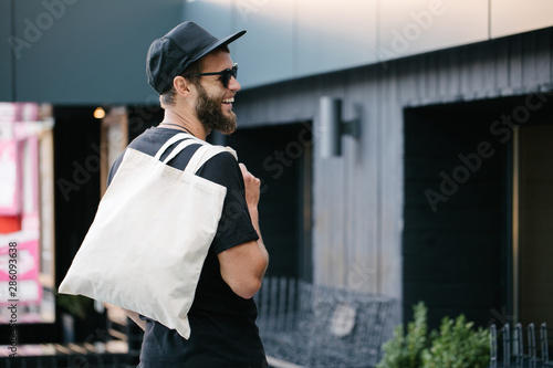 Young man holding white textile eco bag against urban city background. . Ecology or environment protection concept. White eco bag for mock up.