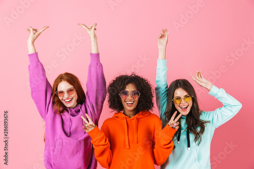 Pleased smiling young three multiethnic girls friends posing isolated over pink wall background showing peace gesture.