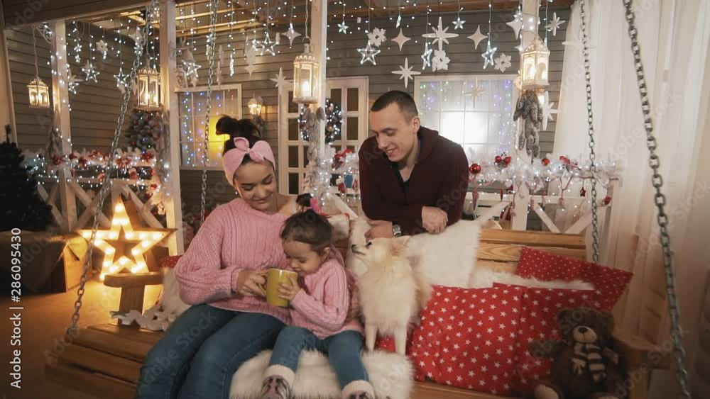 Mother, father, little daughter and white dog have fun sitting at Christmas bench. Young family in pullovers celebrating winter holidays at porch, expressing love and care in slow motion Mother, father, little daughter and white dog have fun sitting at Christmas bench. Young family in pullovers celebrating winter holidays at porch, expressing love and care in slow motion