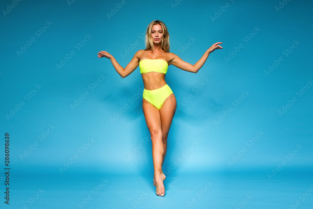young tanned fitness blonde girl in lemony yellow swimsuit is standing like dancer and flirting sexy on the blue wall background, sport lifestyle concept, free space young tanned fitness blonde girl in lemony yellow swimsuit is standing like dancer and flirting sexy on the blue wall background, sport lifestyle concept, free space