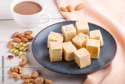 traditional indian candy soan papdi in a blue ceramic plate with almond, pistache and a cup of coffee on a white wooden background with orange textile. side view