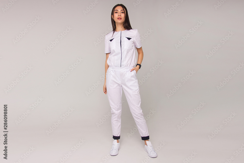 young stylish happy nurse with long brunette hair is standing with hand in her pocket, looking straight and smiling on white wall background. medical fashion concept. free space young stylish happy nurse with long brunette hair is standing with hand in her pocket, looking straight and smiling on white wall background. medical fashion concept. free space