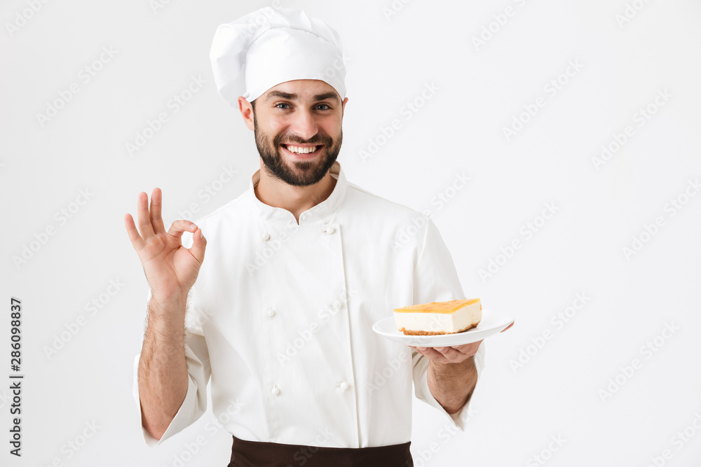 Image of young chief man in cook uniform smiling and showing delicious sign while holding plate with piece of cheesecake Image of young chief man in cook uniform smiling and showing delicious sign while holding plate with piece of cheesecake