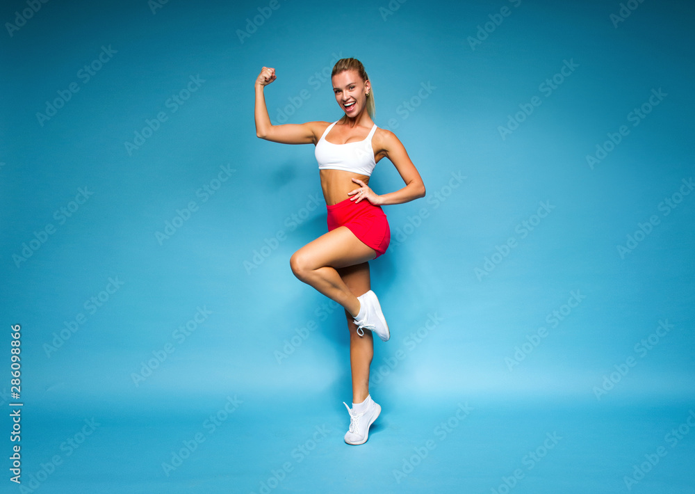 young sports happy sexy blonde girl in red skirt and white top is standing like athlete with strong hands, looking straight and smiling on the blue wall background, sport concept, free space
