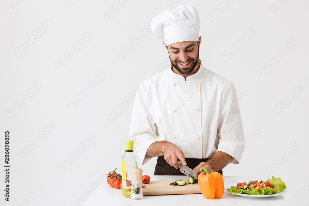 Image of happy chief man in uniform smiling and cooking vegetable salad on wooden cutting board Image of happy chief man in uniform smiling and cooking vegetable salad on wooden cutting board