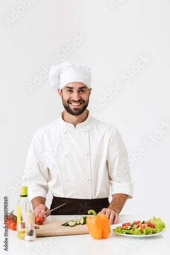 Image of handsome chief man in uniform smiling and cooking vegetable salad on wooden cutting board