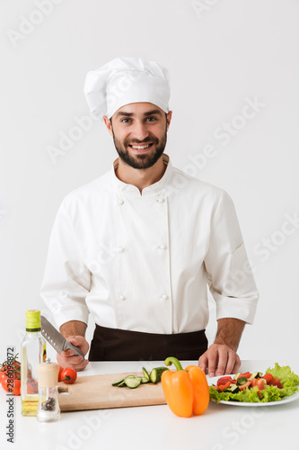 Image of professional chief man in uniform smiling and cooking vegetable salad on wooden cutting board