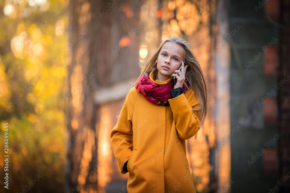 beautiful girl in a bright yellow coat walks in the autumn evening park alone and talks on the phone beautiful girl in a bright yellow coat walks in the autumn evening park alone and talks on the phone