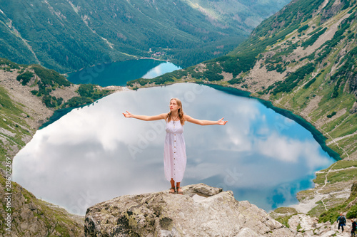 Beautiful long haired girl in white dress standing with hands apart in Polish mountains with fabulous scenic view on background