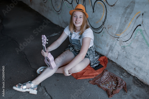 Girl in hat and with pink ukulele sits on the road