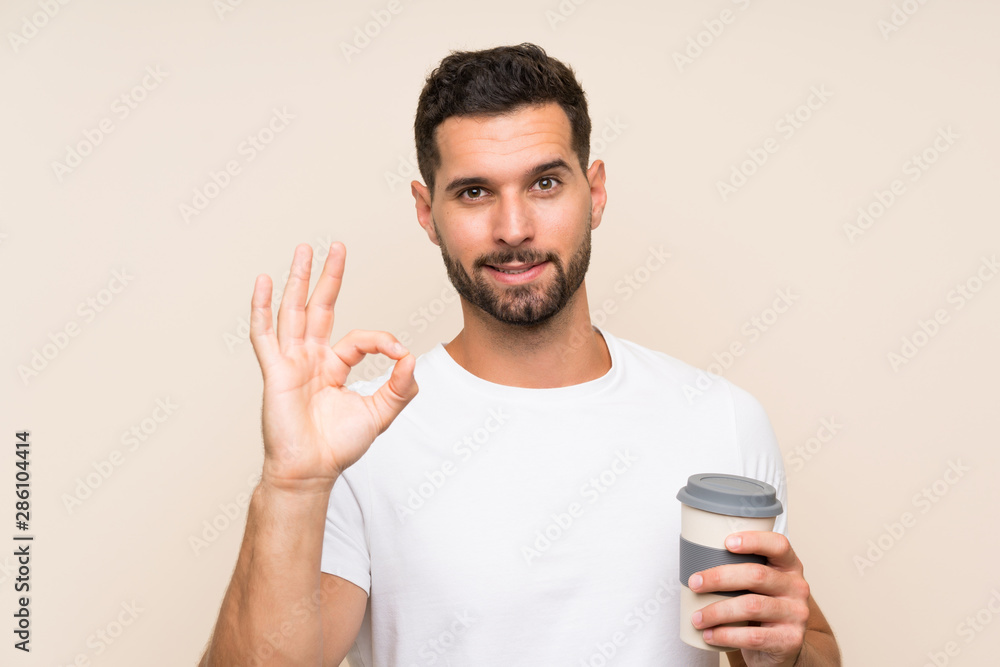 Young man with beard holding a take away coffee over isolated blue background showing ok sign with fingers Young man with beard holding a take away coffee over isolated blue background showing ok sign with fingers