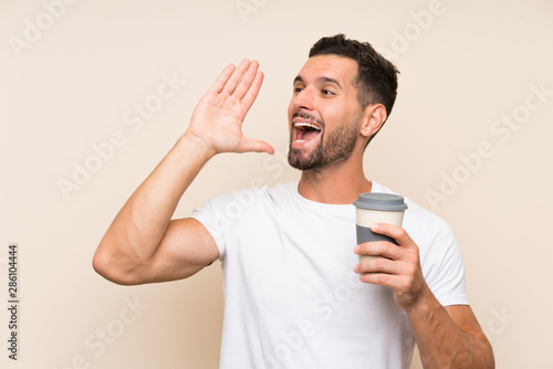 Young man with beard holding a take away coffee over isolated blue background shouting with mouth wide open