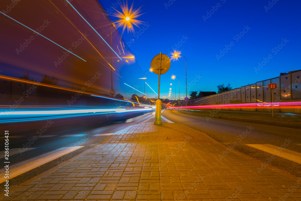 Rozmyty tir ruch drogowy rozmazane światła, na środku znak drogowy i swiecące latarnie - obrazy, fototapety, plakaty 